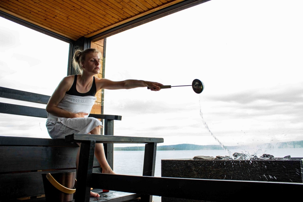 A woman wrapped in a towel pours water on the hot stones in a modern sauna with a glass wall, revealing a vast lake and fell landscape in the background.