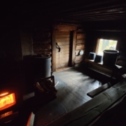 A dimly lit, traditional Finnish log sauna, with the glow of flames flickering through the stove’s hatch and buckets resting on the benches, surrounded by the wilderness of Patvinsuo National Park.
