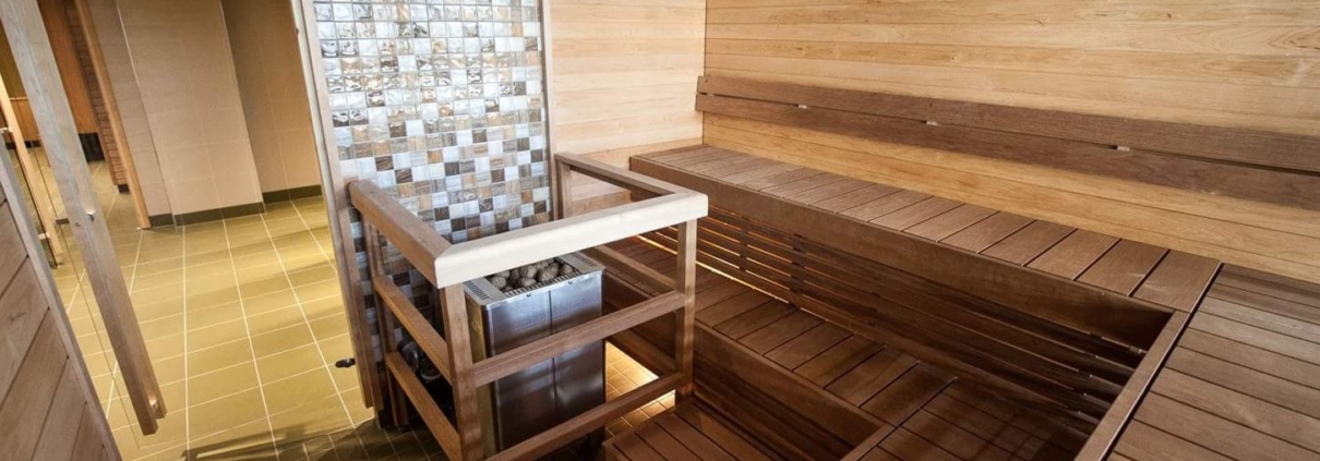 A bright sauna with wooden walls and benches, featuring a centrally placed stove protected by railings.