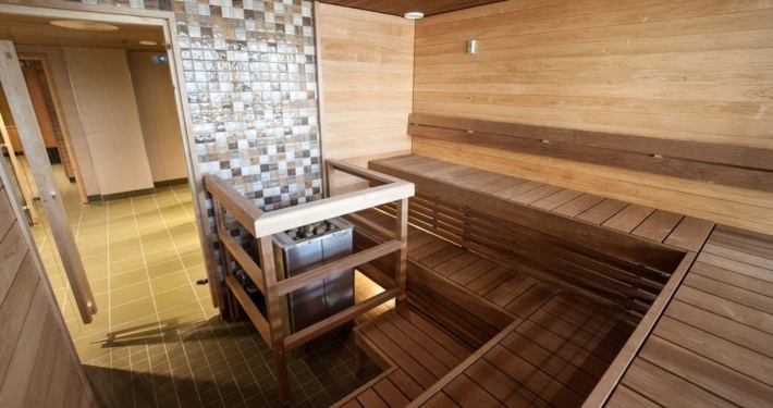 A bright sauna with wooden walls and benches, featuring a centrally placed stove protected by railings.
