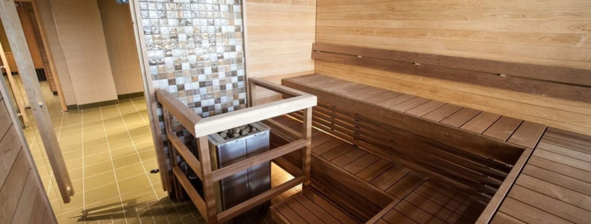 A bright sauna with wooden walls and benches, featuring a centrally placed stove protected by railings.