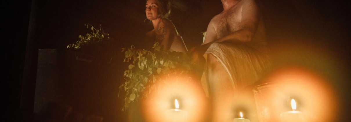 A dimly lit sauna illuminated by candles, where two people are sitting on the upper bench, wrapped in towels, enjoying the warm and relaxing atmosphere with birch whisks nearby.