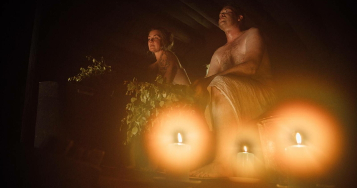 A dimly lit sauna illuminated by candles, where two people are sitting on the upper bench, wrapped in towels, enjoying the warm and relaxing atmosphere with birch whisks nearby.
