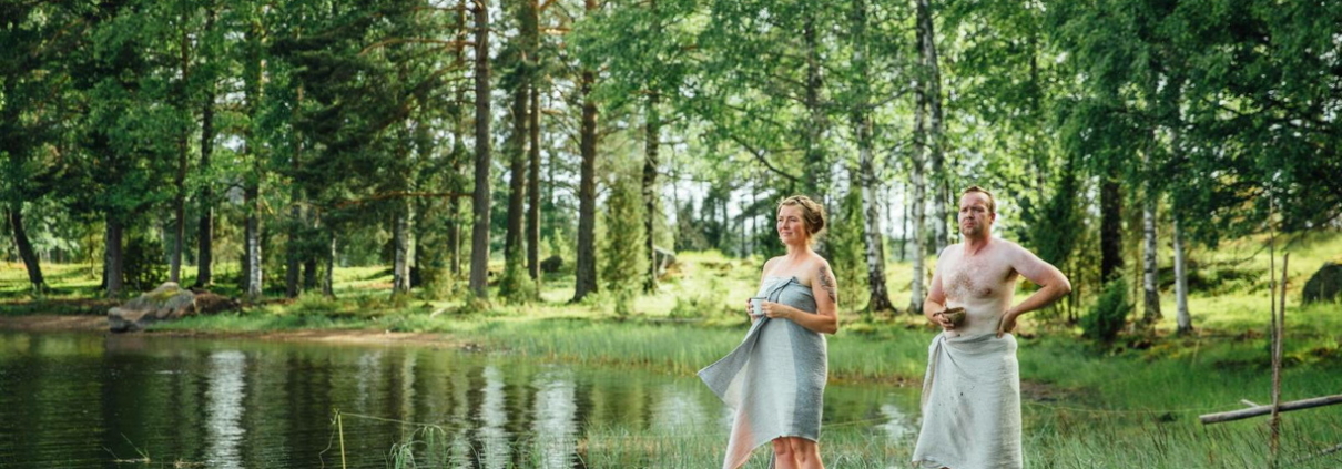A summery Finnish lakeshore with lush green trees, where a man and a woman wrapped in towels stand on a pier, gazing at the lake while cooling off between sauna sessions.