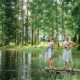 A summery Finnish lakeshore with lush green trees, where a man and a woman wrapped in towels stand on a pier, gazing at the lake while cooling off between sauna sessions.