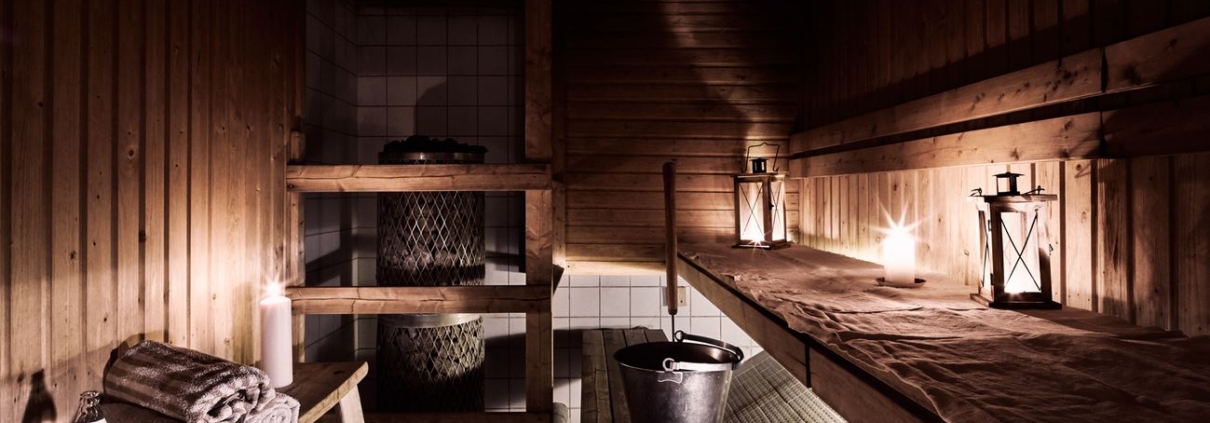 A sauna illuminated atmospherically by lanterns and candles, with walls and benches made of beautiful brown wood. On the benches, there are towels, a bottle of water, a water bucket, and a ladle for throwing steam.