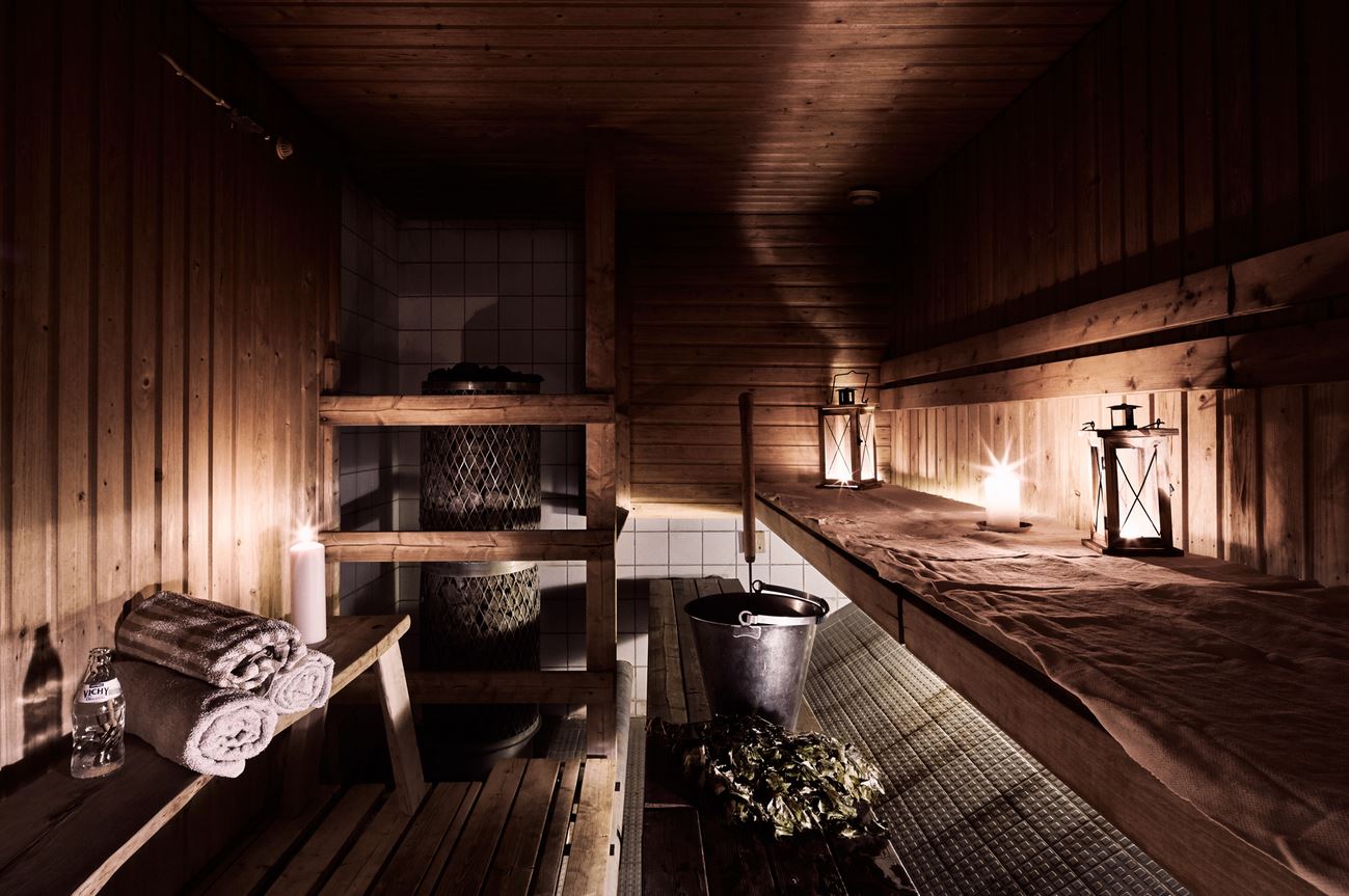 A sauna illuminated atmospherically by lanterns and candles, with walls and benches made of beautiful brown wood. On the benches, there are towels, a bottle of water, a water bucket, and a ladle for throwing steam.