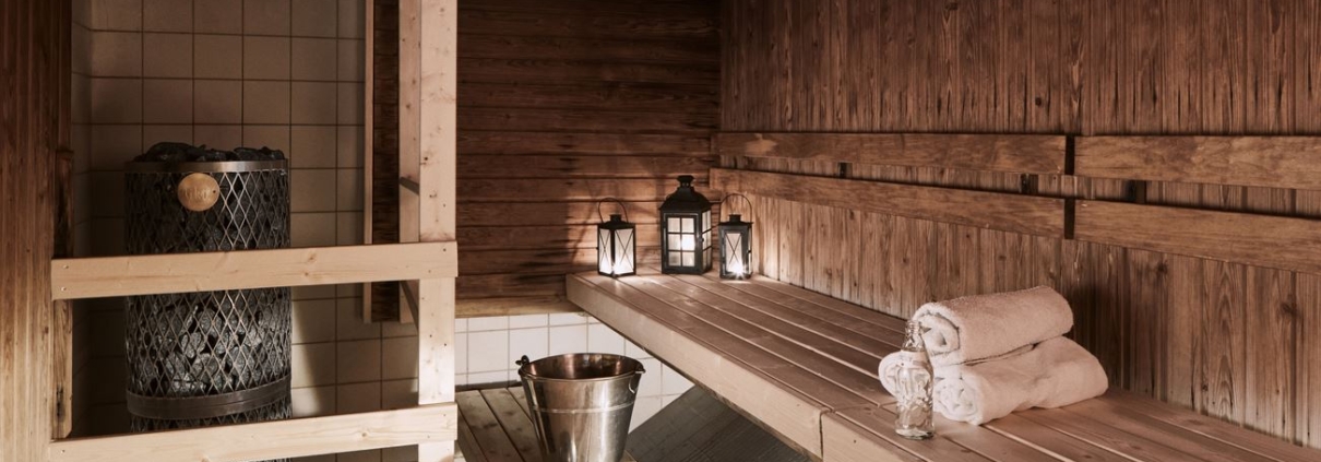 A sauna illuminated atmospherically by lanterns, with walls and benches made of beautiful brown wood. On the benches, there are towels, a bottle of water, a water bucket, and a ladle for throwing steam.