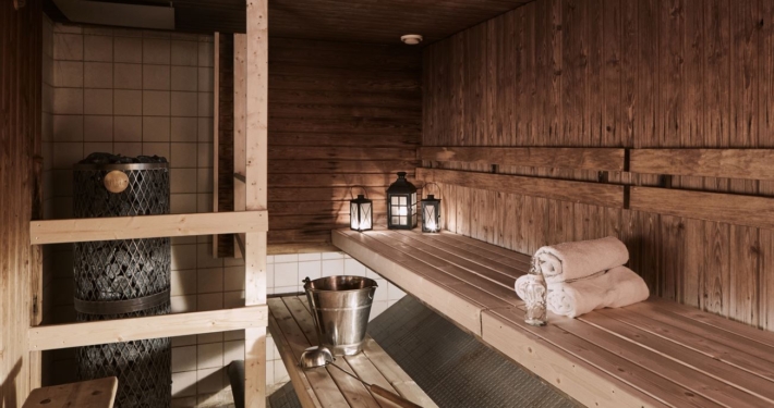 A sauna illuminated atmospherically by lanterns, with walls and benches made of beautiful brown wood. On the benches, there are towels, a bottle of water, a water bucket, and a ladle for throwing steam.