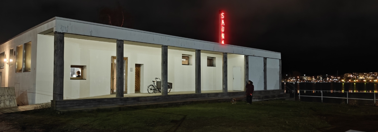 Kulttuurisauna is a white, box-shaped building, with the sea and the lights of Helsinki glowing in the dark evening background.