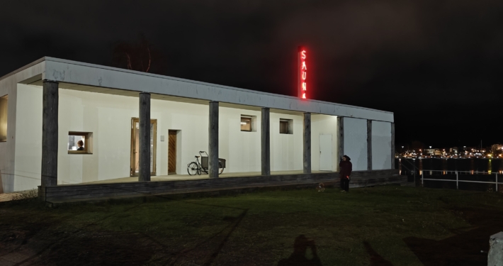 Kulttuurisauna is a white, box-shaped building, with the sea and the lights of Helsinki glowing in the dark evening background.
