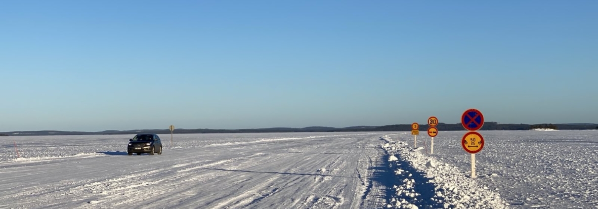 Under the blue sky stretches a white, snow-covered ice field—a frozen lake with an ice road. A car is driving on the ice road, and several traffic signs are visible.