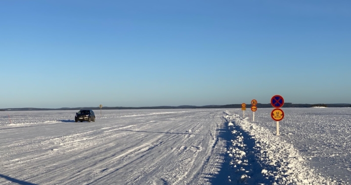 Under the blue sky stretches a white, snow-covered ice field—a frozen lake with an ice road. A car is driving on the ice road, and several traffic signs are visible.