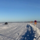 Under the blue sky stretches a white, snow-covered ice field—a frozen lake with an ice road. A car is driving on the ice road, and several traffic signs are visible.