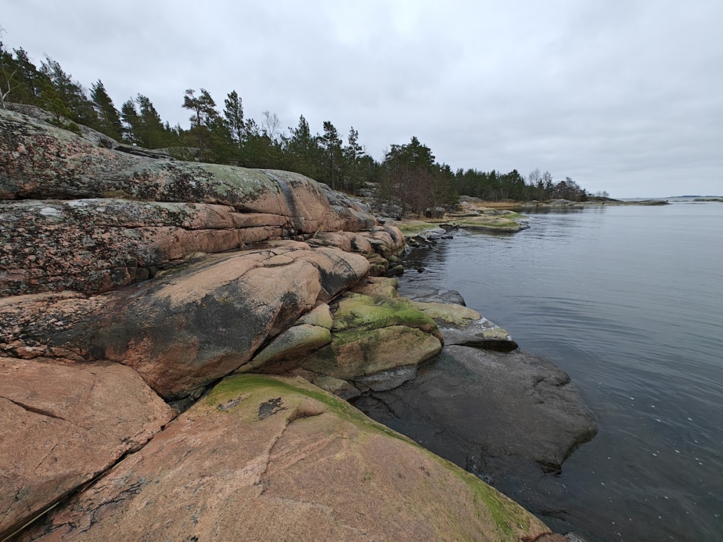 Pinkish and gray smooth rocks on a forested seashore, the sea is calm and peaceful, and the sky is gray.