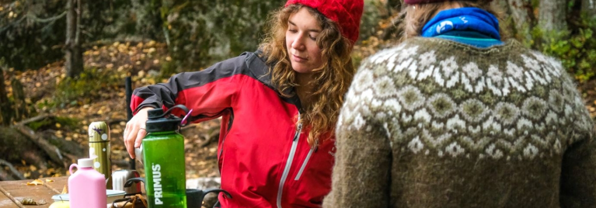 Two people are sitting at a picnic table in an autumn forest and have laid out their food on the table.