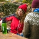 Two people are sitting at a picnic table in an autumn forest and have laid out their food on the table.