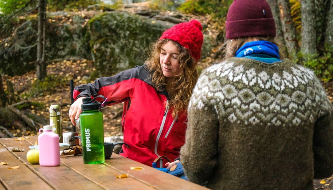 Two people are sitting at a picnic table in an autumn forest and have laid out their food on the table.